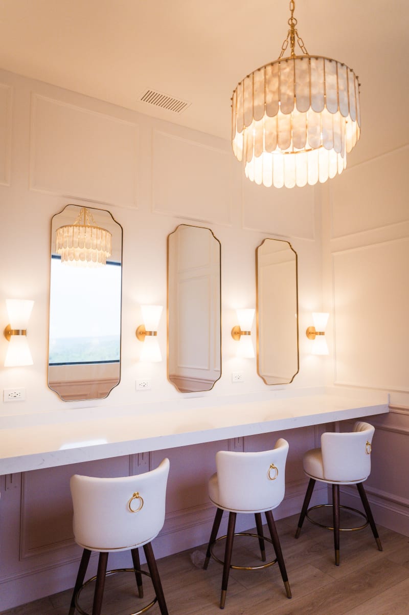 Bridal suite vanity at The Chapel at Wimberley with gold-framed mirrors, sconces, a capiz chandelier, and white swivel stools