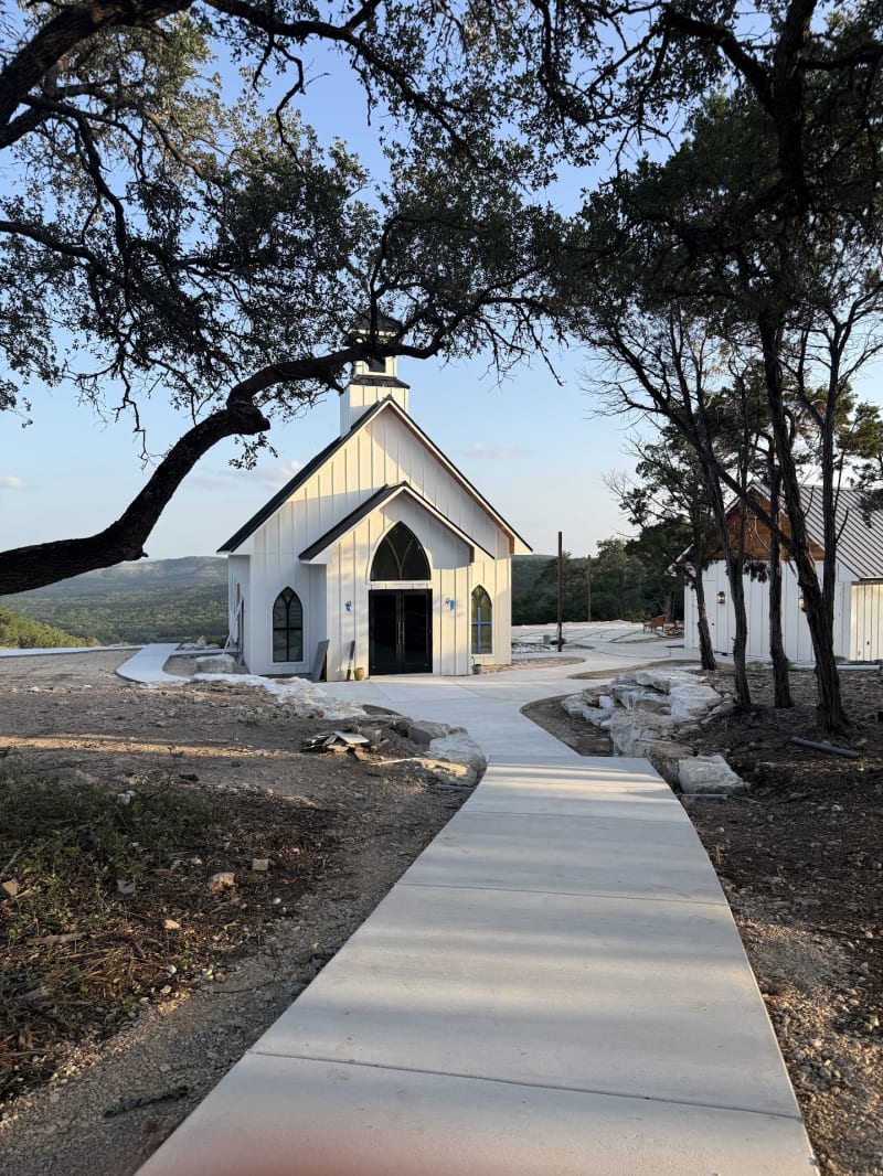 A walkway beneath oak trees leading to the white chapel in golden evening light